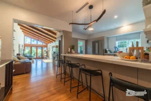 Open living area with modern kitchen, breakfast bar, and vaulted wooden ceiling by Ground Up Builders