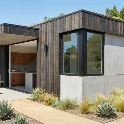 A modern, boxy accessory dwelling unit (ADU) featuring vertical dark wood siding mixed with concrete walls, a flat roof, and large corner windows facing a xeriscaped garden.