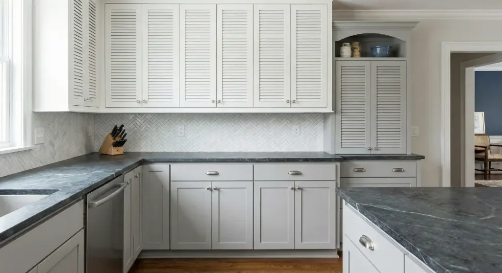 Light grey kitchen cabinets with louvered upper doors and dark grey stone countertops.